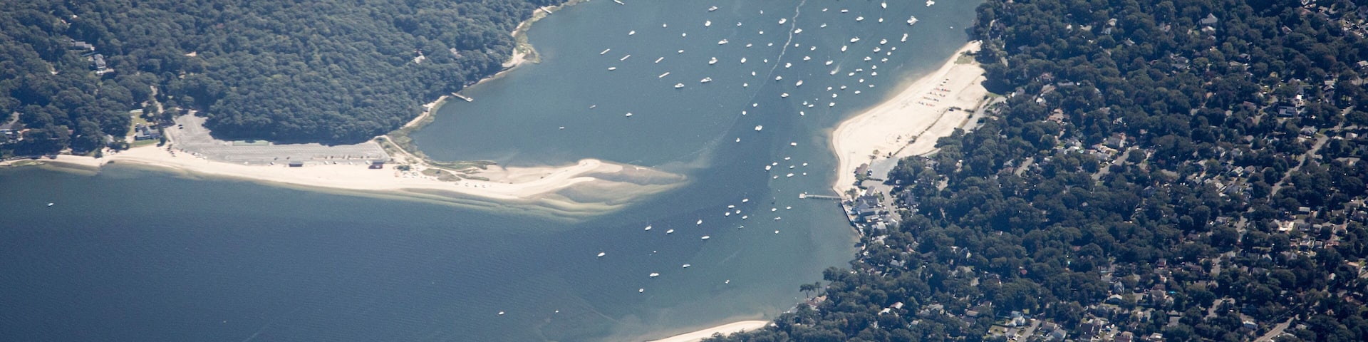 An aerial view of a coastal area with a sandy beach, dense forest, and a marina with numerous boats.