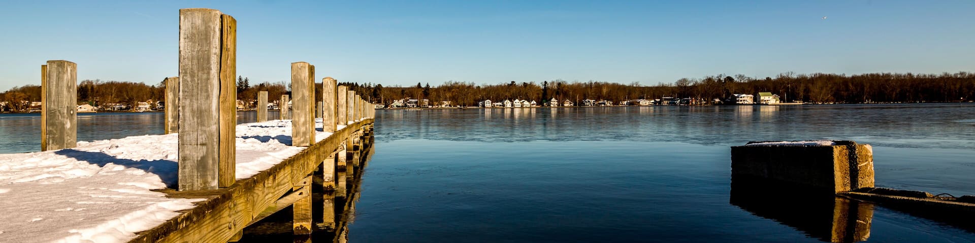A wooden pier extending into a calm lake with snow on the ground, surrounded by trees and houses in the background.