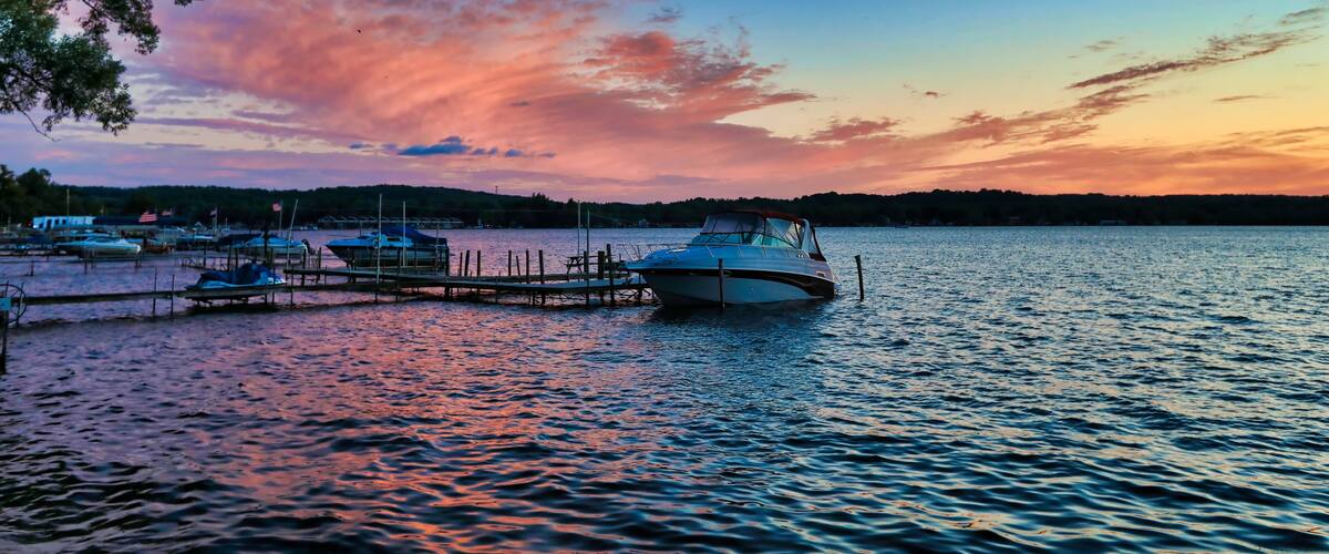 A calm lake at sunset with boats docked at a pier and a forested shoreline in the background.
