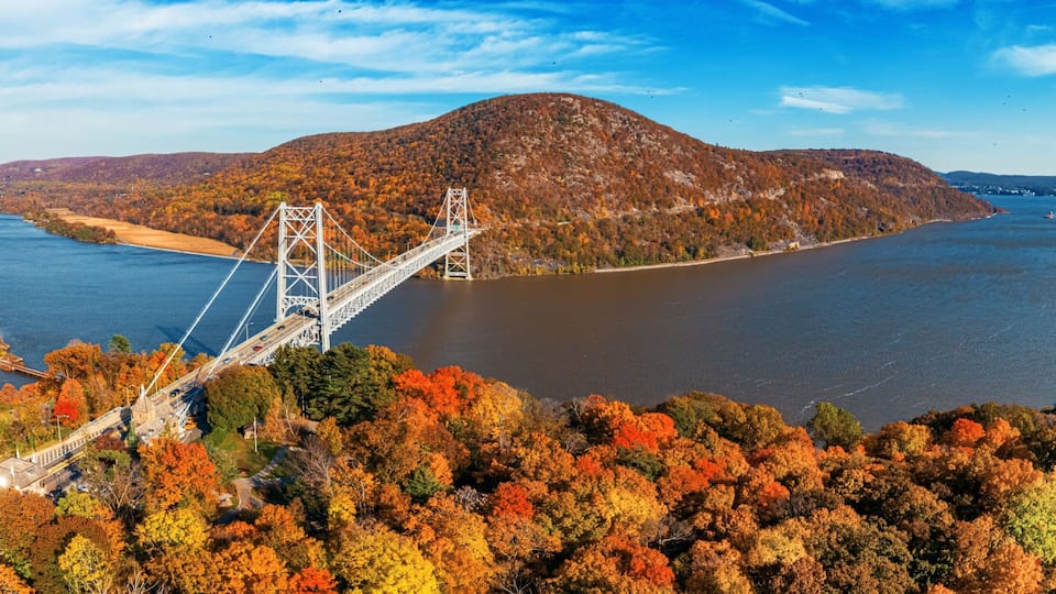 A suspension bridge over a river, surrounded by autumn-colored trees and hills.