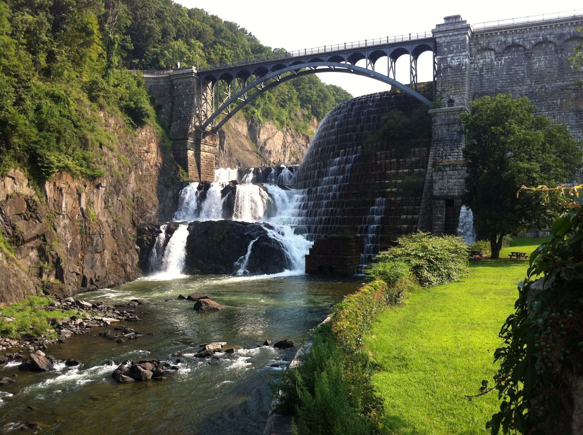 A stone bridge over a river with multiple waterfalls, surrounded by lush greenery and a picnic area.