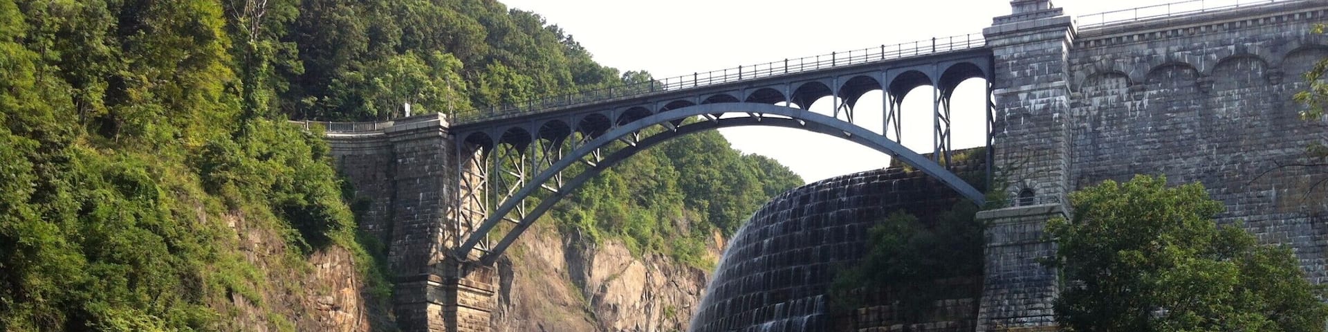 A stone bridge over a river with multiple waterfalls, surrounded by lush greenery and a picnic area.