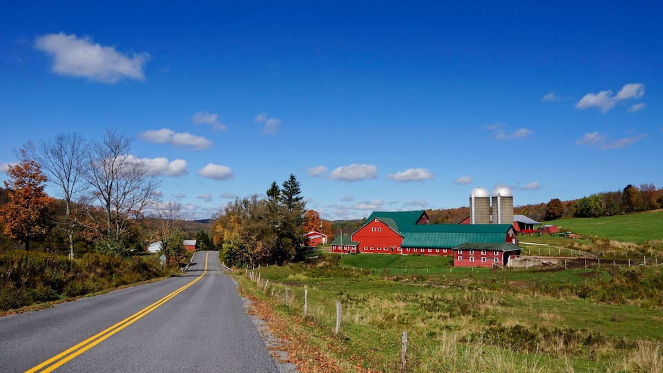 A rural road with a red barn and silos, surrounded by autumn foliage and a clear blue sky.