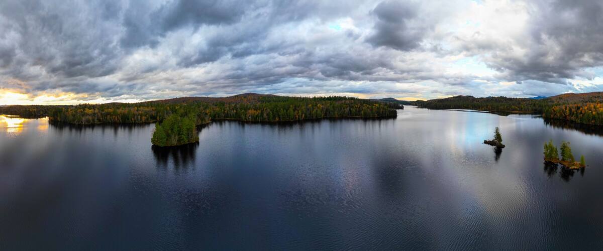 A calm lake surrounded by dense forests with autumn foliage, under a cloudy sky.