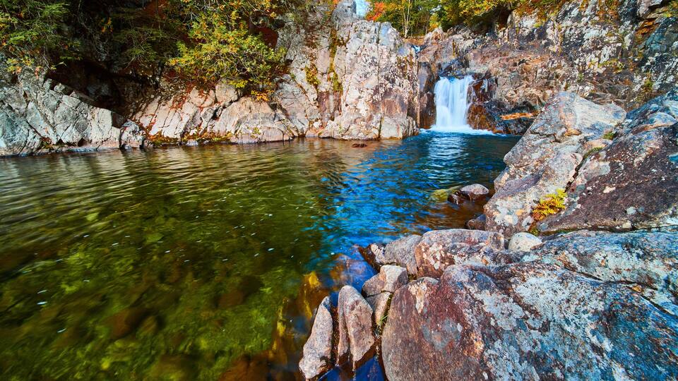 Small waterfall over rocks into blue and green river next to huge boulders in forest