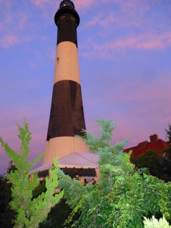 A lighthouse with a white and black striped tower, surrounded by greenery and a red building in the background.