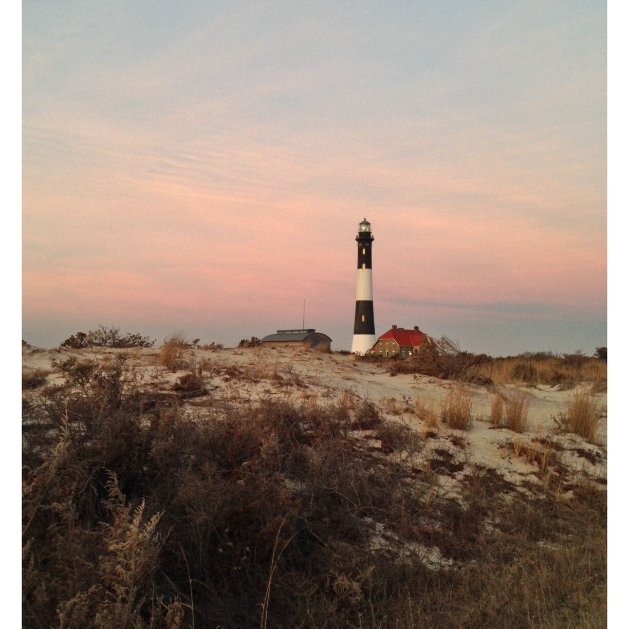 A lighthouse on a sandy dune with sparse vegetation and a red-roofed building nearby.