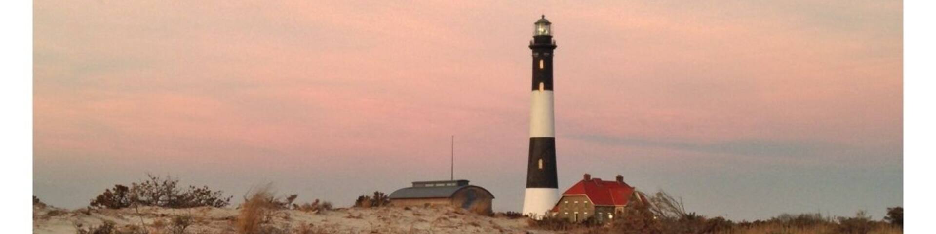 A lighthouse on a sandy dune with sparse vegetation and a red-roofed building nearby.