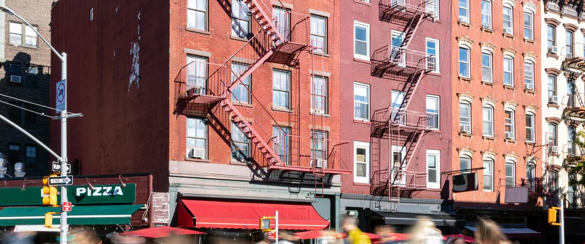 A busy street with pedestrians crossing, a pizza restaurant, and red brick buildings with fire escapes.