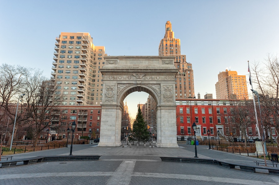 Um arco com esculturas detalhadas, rodeado por edifícios altos e árvores sem folhas.