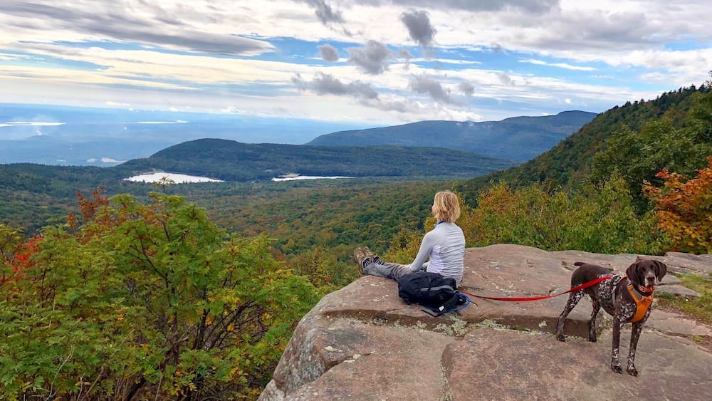A person sitting on a rock overlooking a mountainous landscape with a lake and autumn foliage.