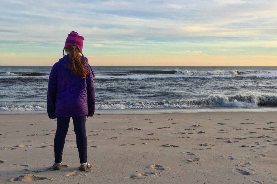A person standing on a sandy beach with footprints, looking out at the ocean with waves.