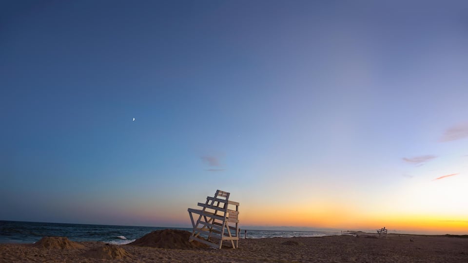 A beach with a lifeguard chair and a sign, ocean waves, and a sunset sky.