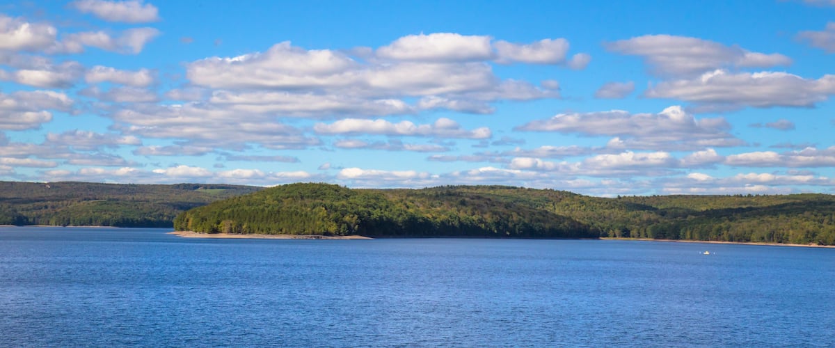 A calm lake with a forested island and a clear blue sky with scattered clouds.
