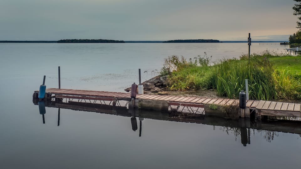 A calm lake with a wooden dock extending into the water, surrounded by grassy banks and a distant shoreline.
