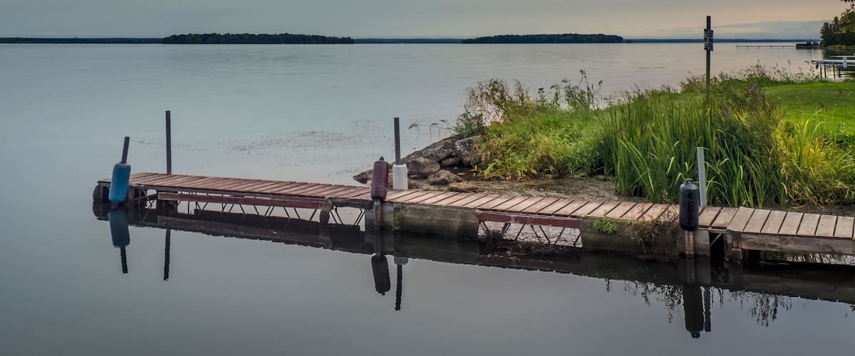 A calm lake with a wooden dock extending into the water, surrounded by grassy banks and a distant shoreline.