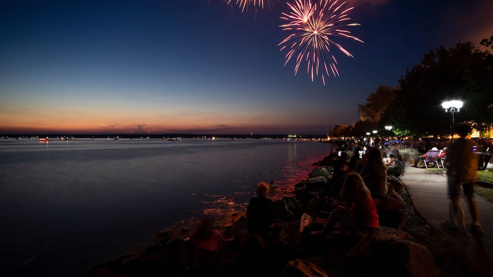 A waterfront with fireworks in the sky, a calm body of water, and people sitting on rocks.