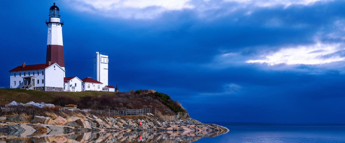 A lighthouse with a red top, white base, and a small house beside it, overlooking a calm body of water with a rocky shoreline.