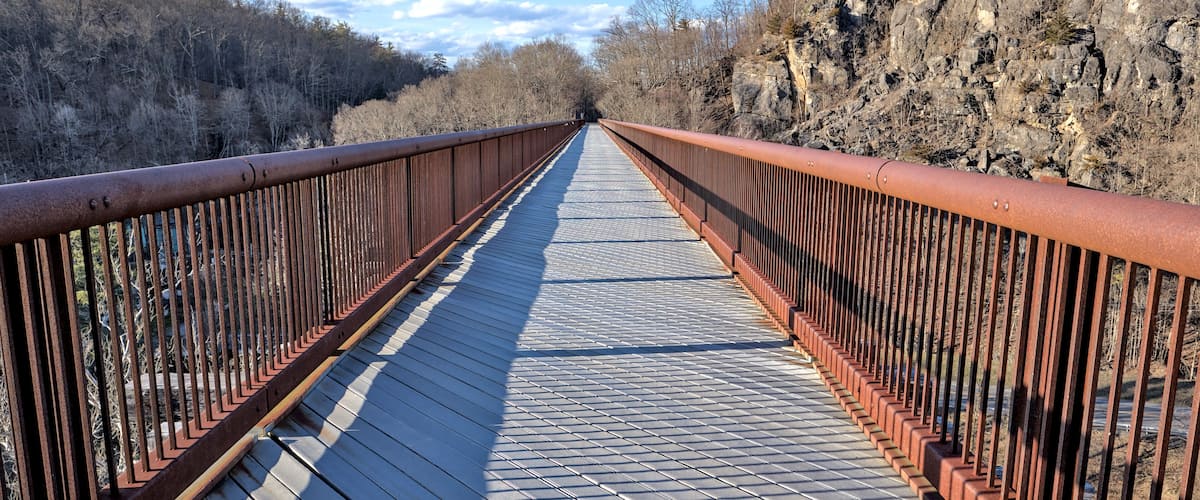 A wooden bridge with metal railings, surrounded by rocky terrain and sparse trees.
