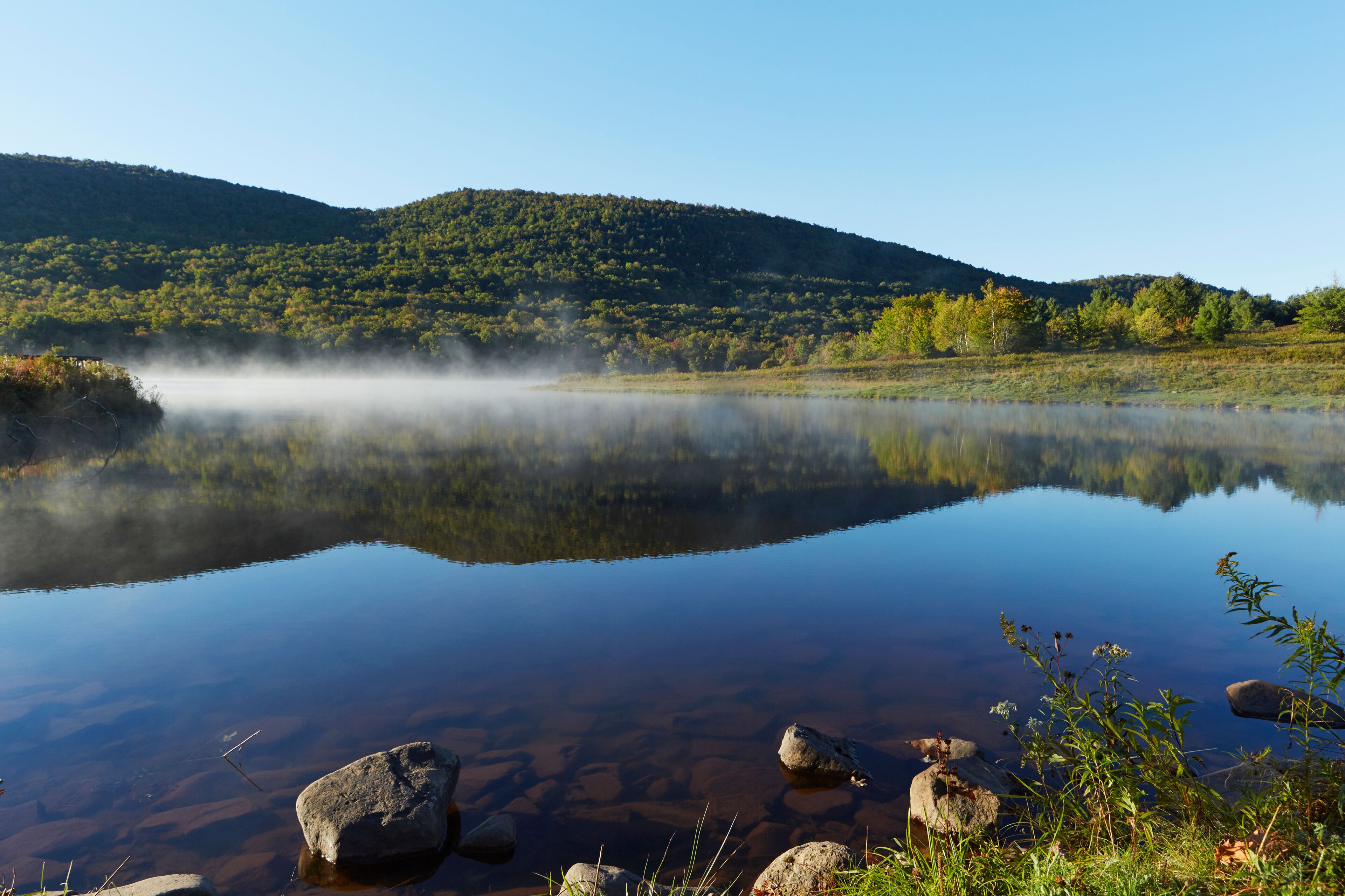 A calm lake with clear reflections, surrounded by green hills and rocks.