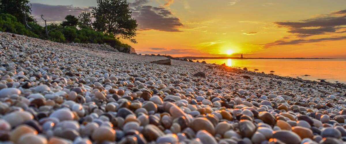 A pebbled beach with a sunset and a distant bridge.