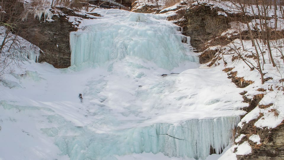 A frozen waterfall surrounded by snow-covered terrain and leafless trees.