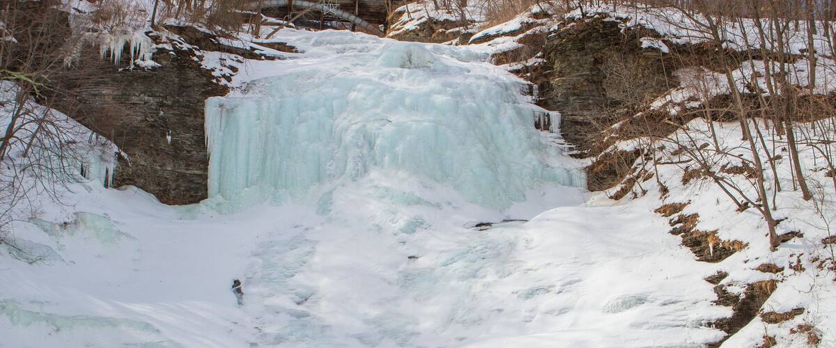 A frozen waterfall surrounded by snow-covered terrain and leafless trees.