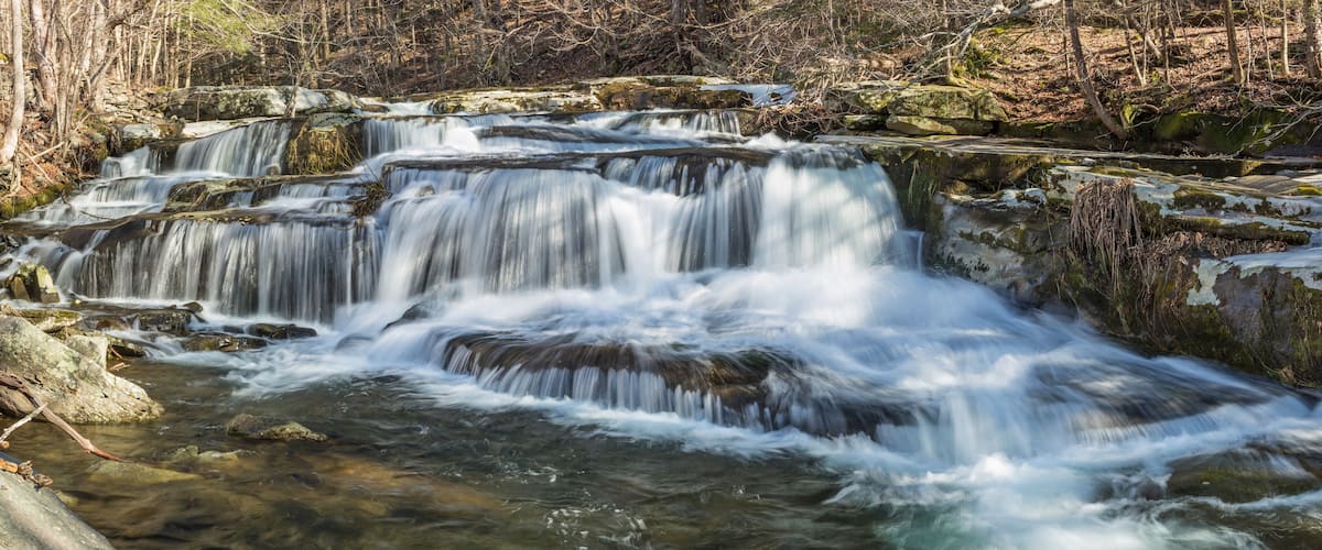 A multi-tiered waterfall surrounded by rocky terrain and sparse trees.
