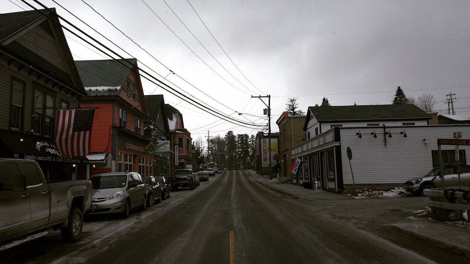 A snow-covered street with parked cars and buildings, including a shop with an American flag.
