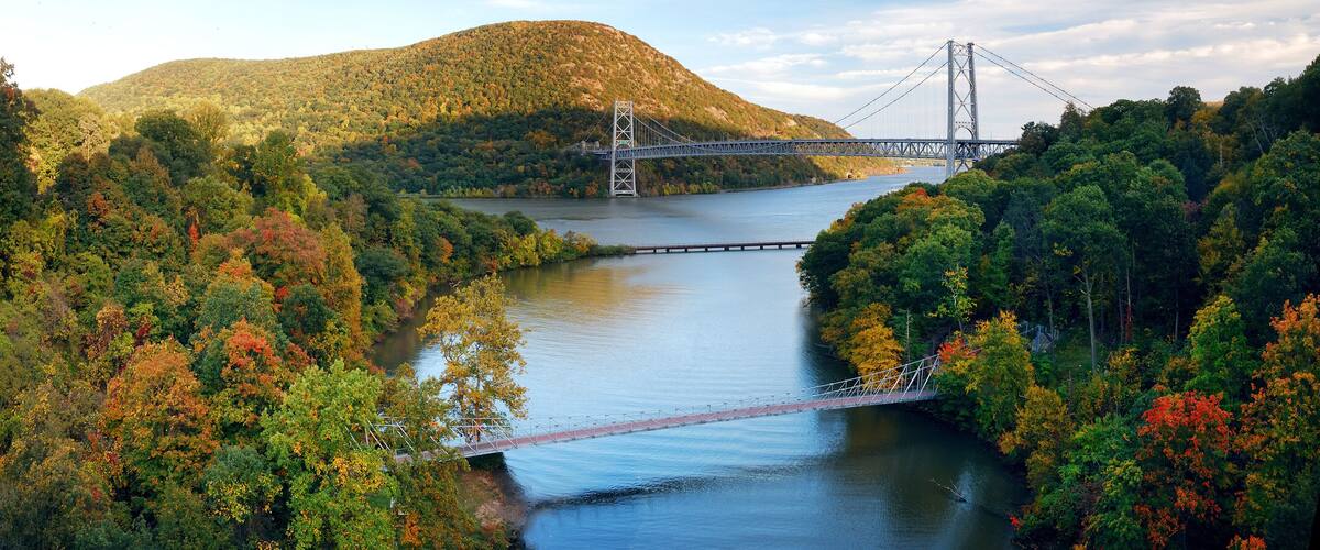 A suspension bridge over a river surrounded by autumn foliage and hills.