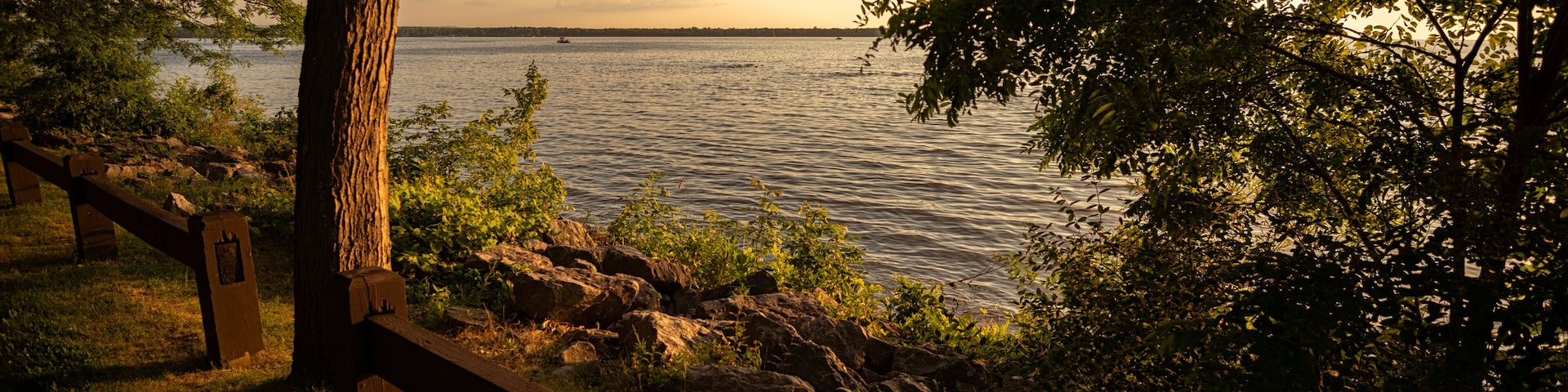 Vista sobre un lago con una cerca de madera, árboles y un barco distante en el agua.