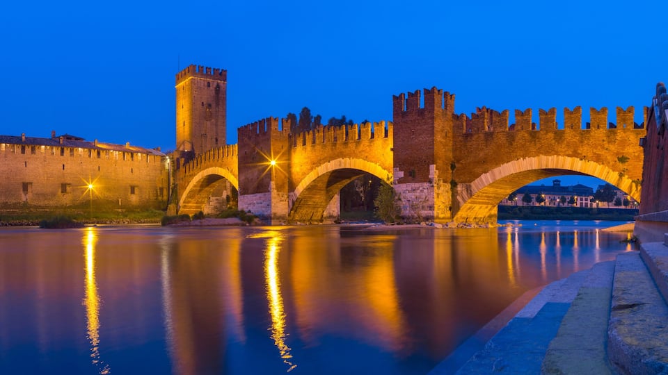 A historic stone bridge with arches over a calm river, surrounded by medieval walls and a tower.