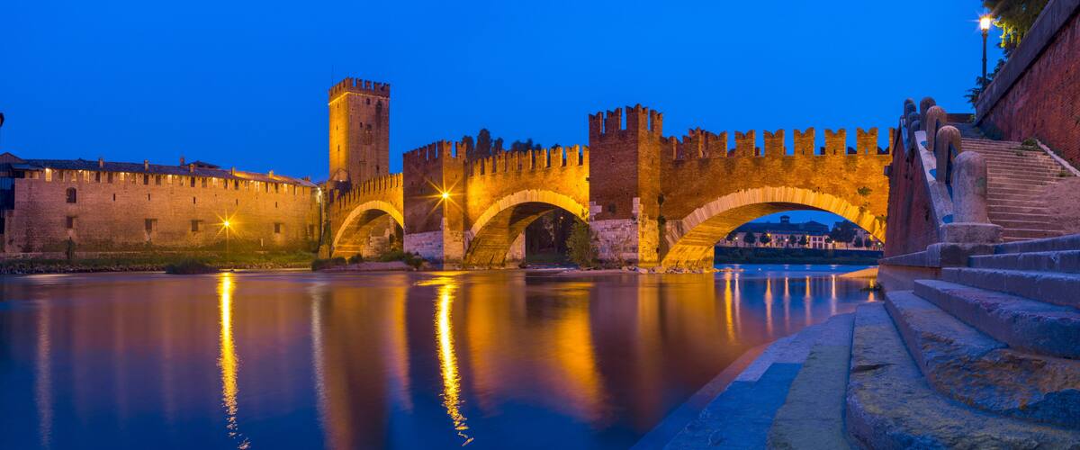 A historic stone bridge with arches over a calm river, surrounded by medieval walls and a tower.
