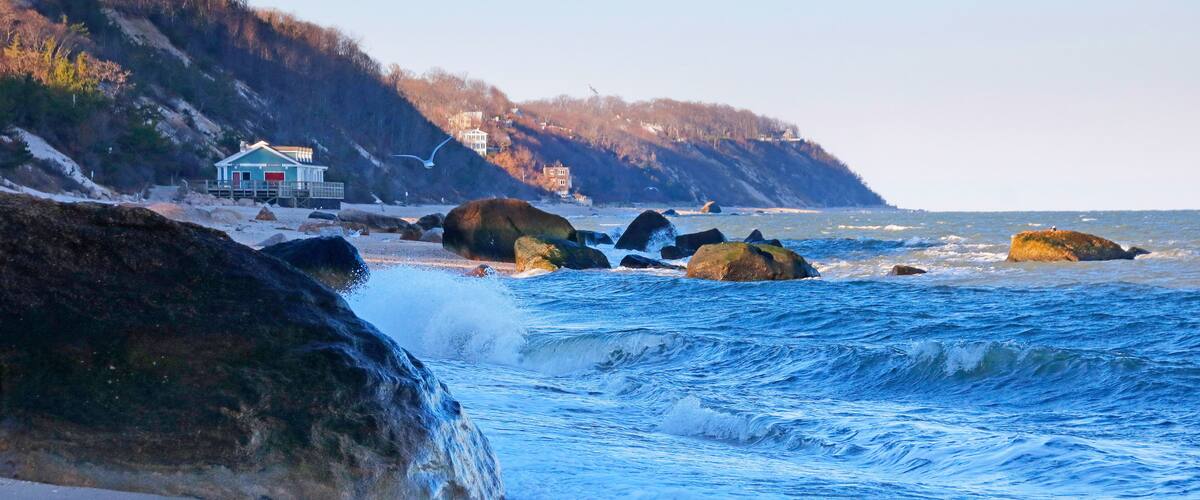 A rocky shoreline with waves crashing onto the beach, a house on stilts, and a forested hillside in the background.