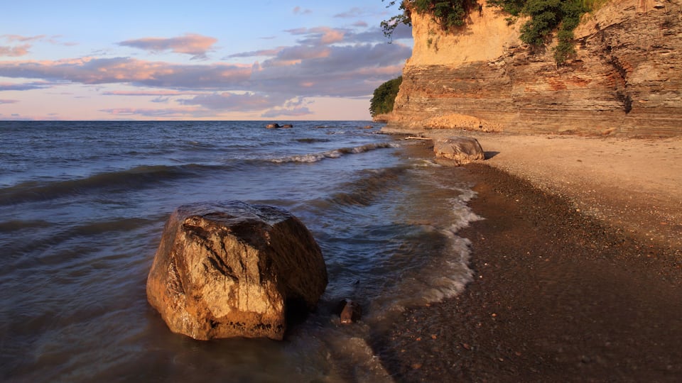 A rocky beach with waves, a large boulder, and a cliff with vegetation.