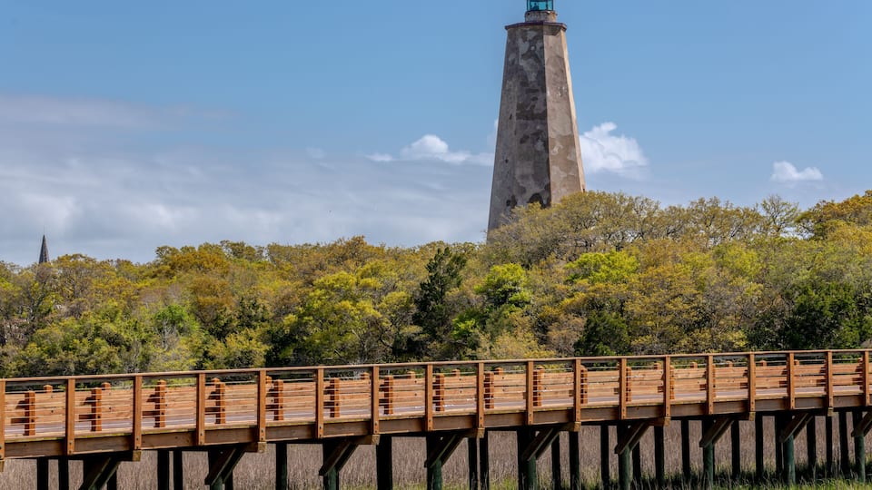 A wooden bridge leading to a tall lighthouse with a weathered tower, surrounded by trees and a clear sky.