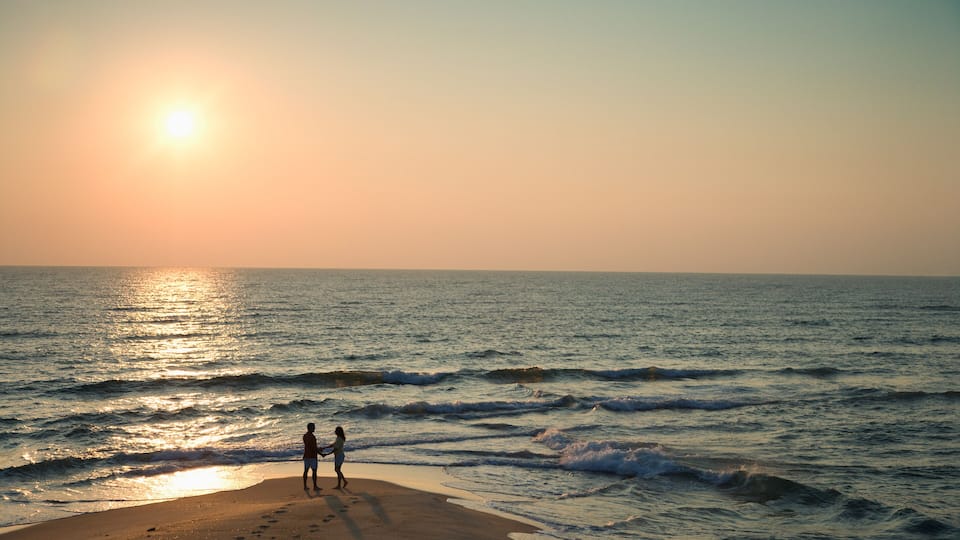 A beach with two people standing near the water's edge during sunset.