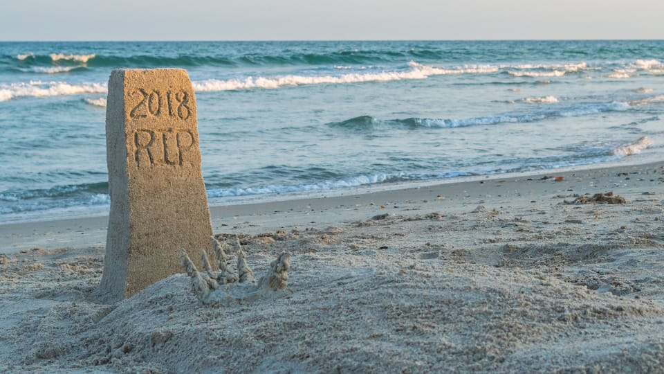 A sand marker with '2018 RIP' inscribed, a sandy beach, and waves in the background.