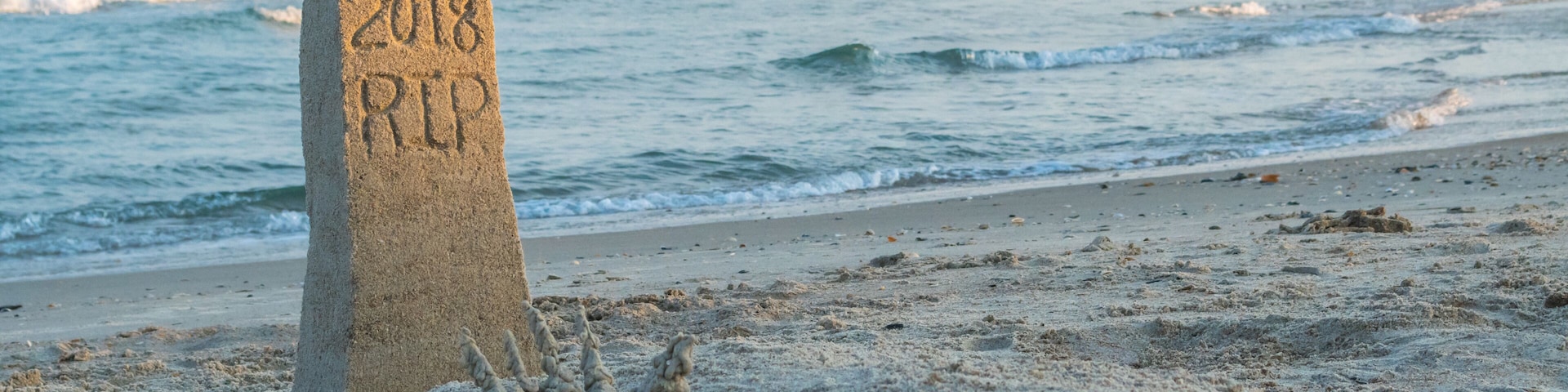 A sand marker with '2018 RIP' inscribed, a sandy beach, and waves in the background.
