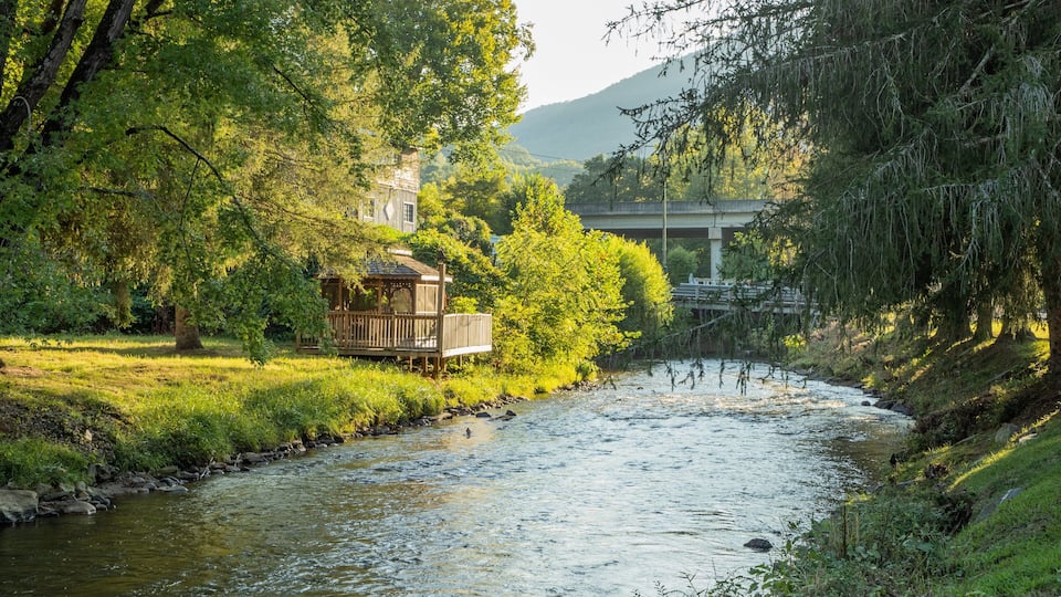 A river with a wooden structure on the bank, surrounded by trees and a bridge in the background.