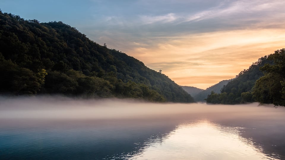 A calm lake surrounded by forested hills during sunrise or sunset.