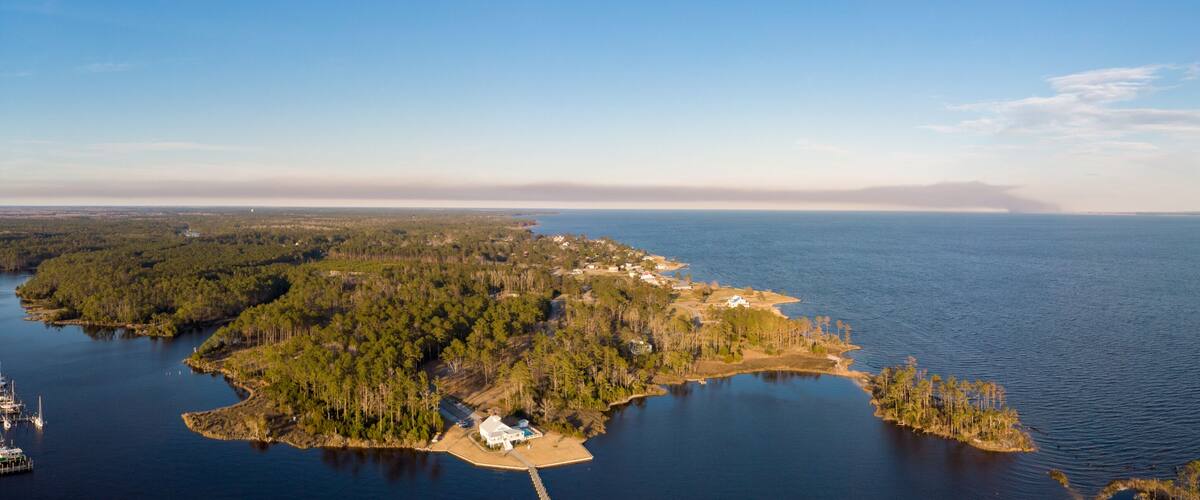 An aerial view of a coastal area with a small island surrounded by water, featuring dense forests, a marina, and a few buildings.