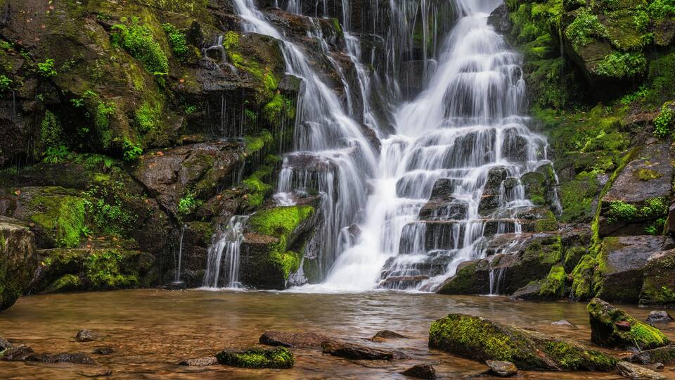 A multi-tiered waterfall surrounded by moss-covered rocks and a calm pool at the base.