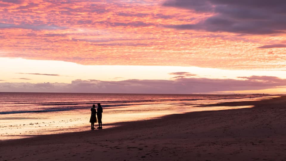 A beach with a sunset sky and two silhouetted figures.