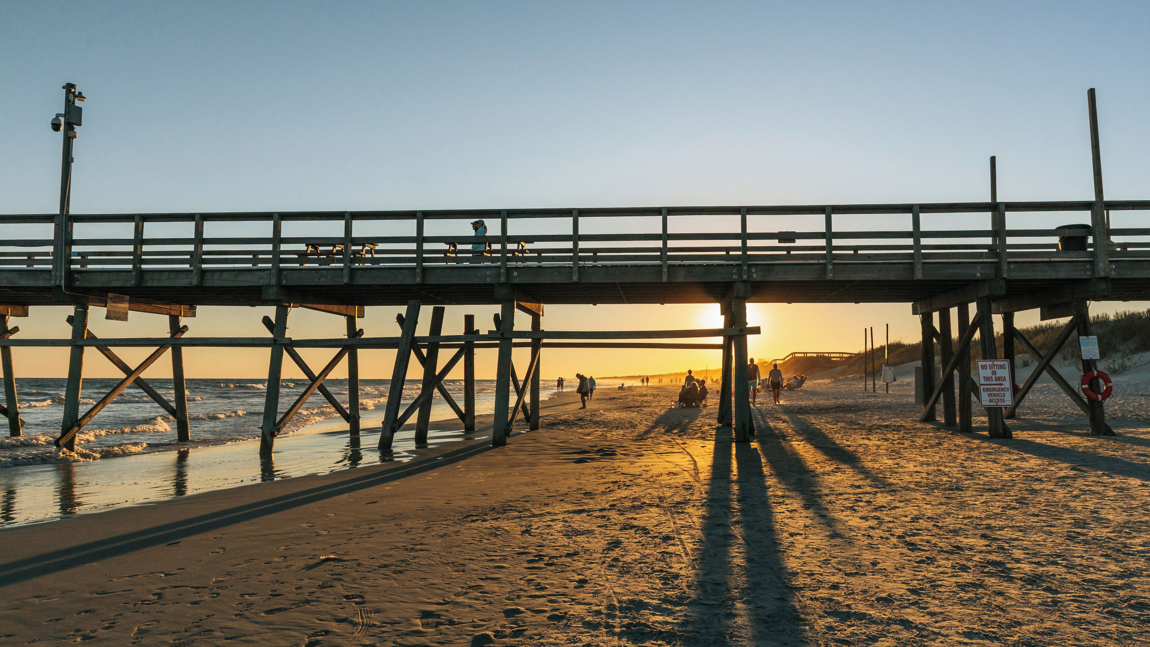 A wooden pier extending over a sandy beach with footprints leading to the water, a lifebuoy, and a sign prohibiting sitting in the area.
