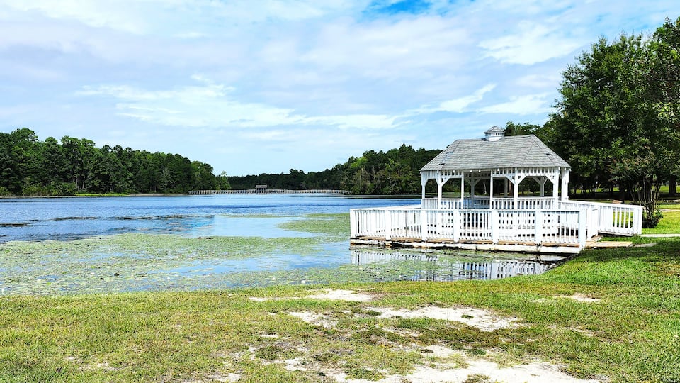 A gazebo by a lake with a dock, surrounded by trees and grassy areas.