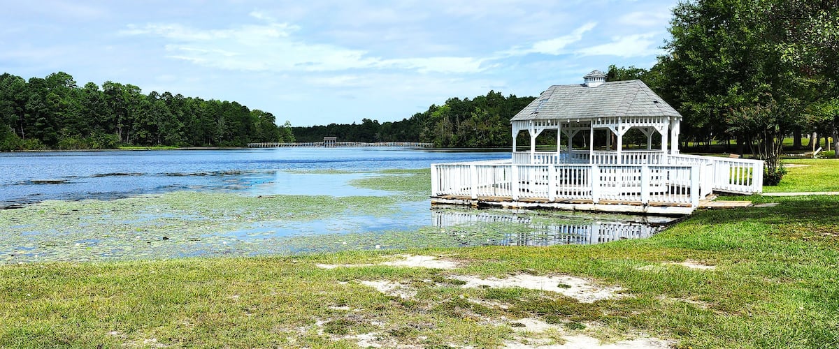 A gazebo by a lake with a dock, surrounded by trees and grassy areas.