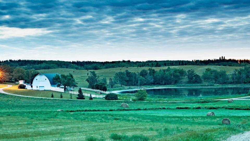A rural landscape with a winding road, hay bales, a white barn, and a house with a porch.