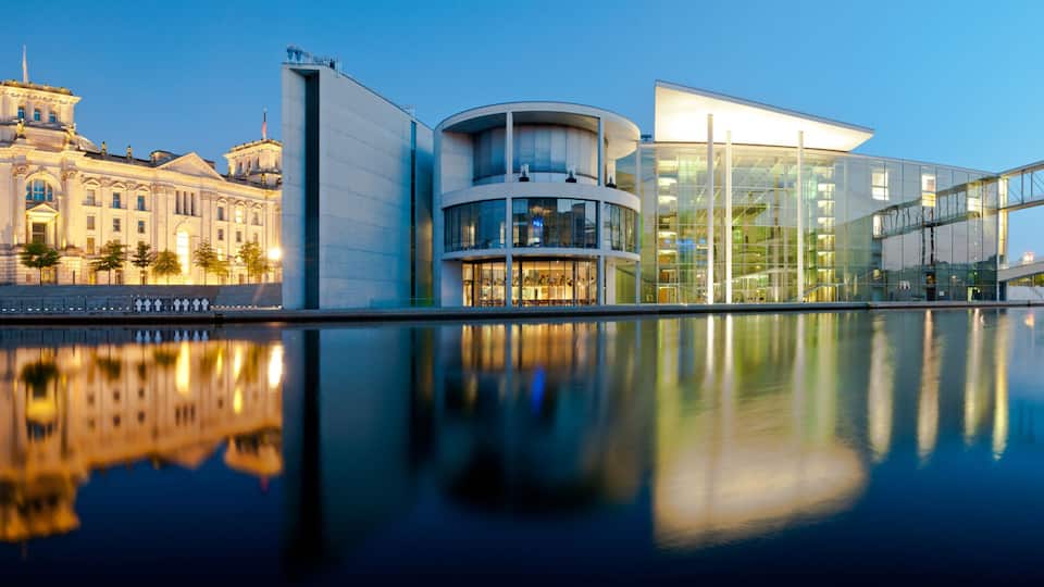 A waterfront with a classical building, a modern glass structure, and a bridge.
