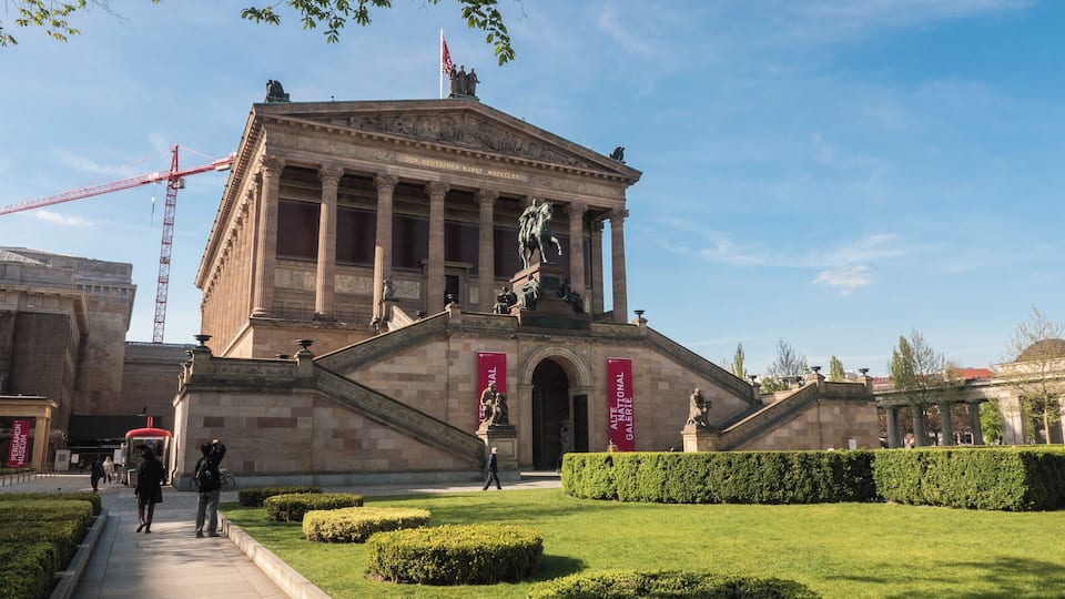A classical building with columns and statues, surrounded by well-manicured lawns and a clear sky.